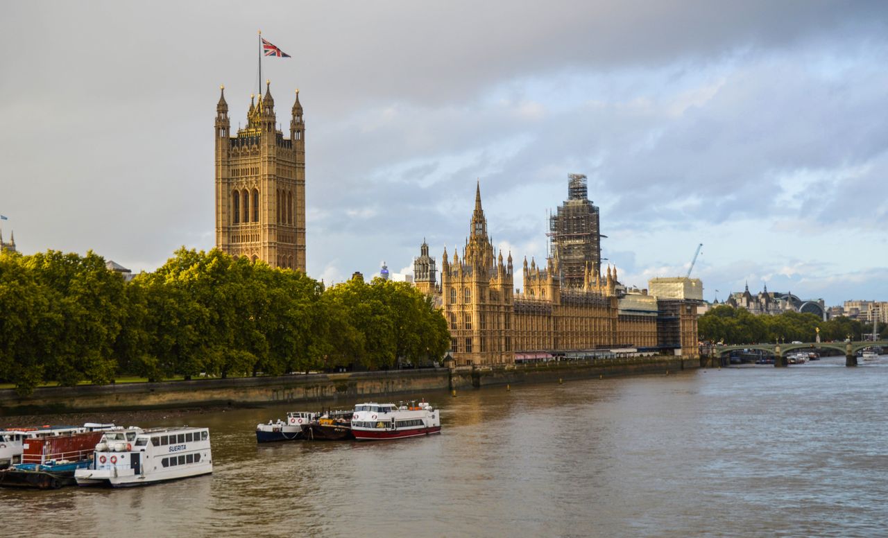 Exterior of aged building of the Houses of Parliament on bushy coast near river with vessels under cloudy sky of London